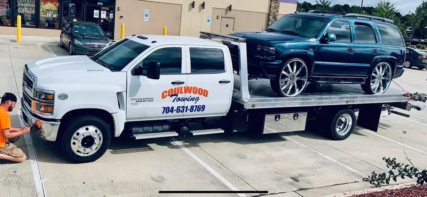 A flatbed tow truck carrying a dark teal SUV with large chrome wheels, parked in a lot with a worker nearby.