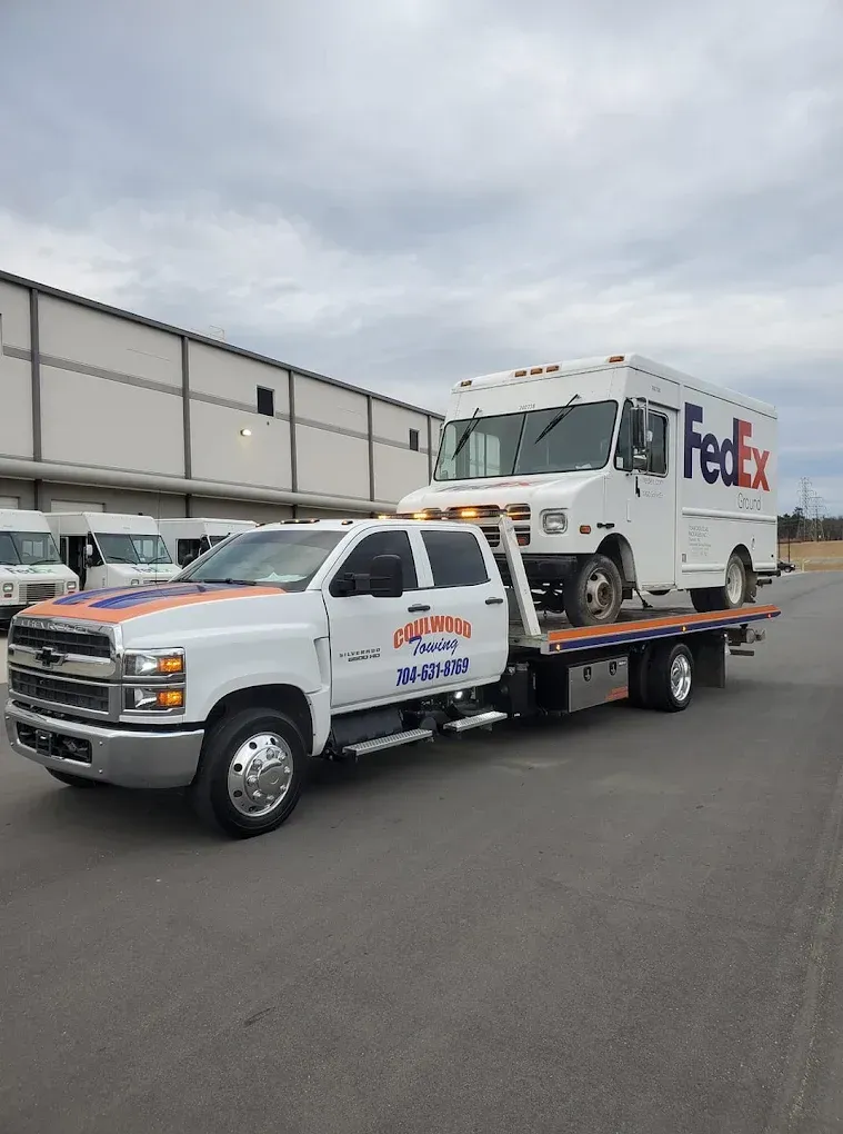 A white FedEx delivery truck being hauled on the flatbed of a white Chevrolet tow truck in an industrial parking lot.