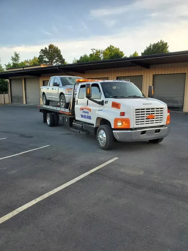 A white flatbed tow truck carrying a silver pickup truck parked in front of a commercial storage facility.