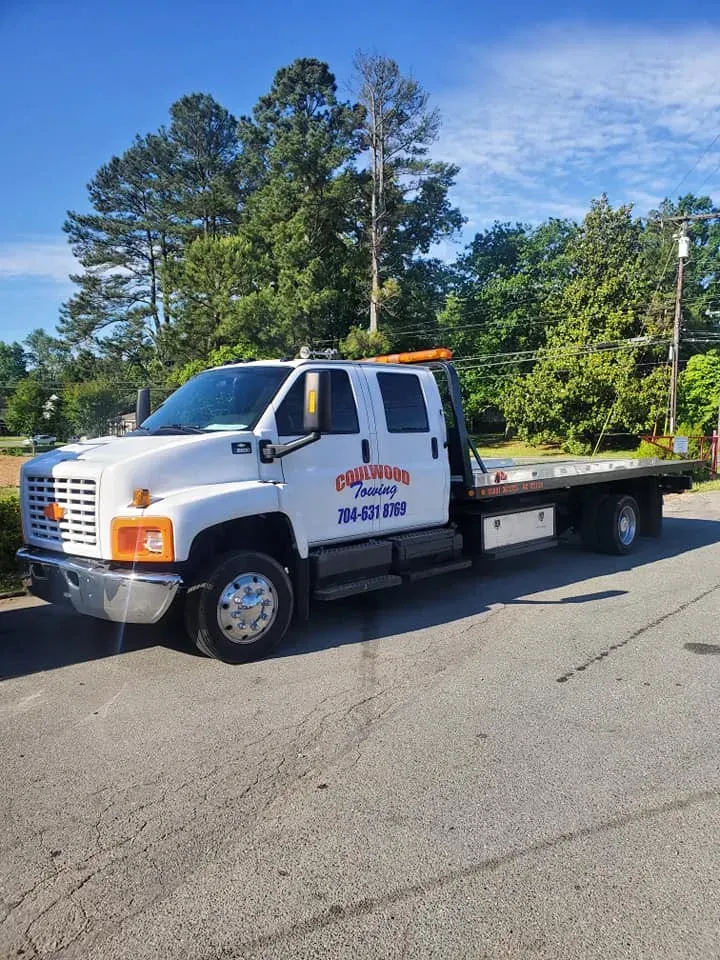A white crew-cab rollback tow truck parked on an asphalt road in front of trees under a clear blue sky.
