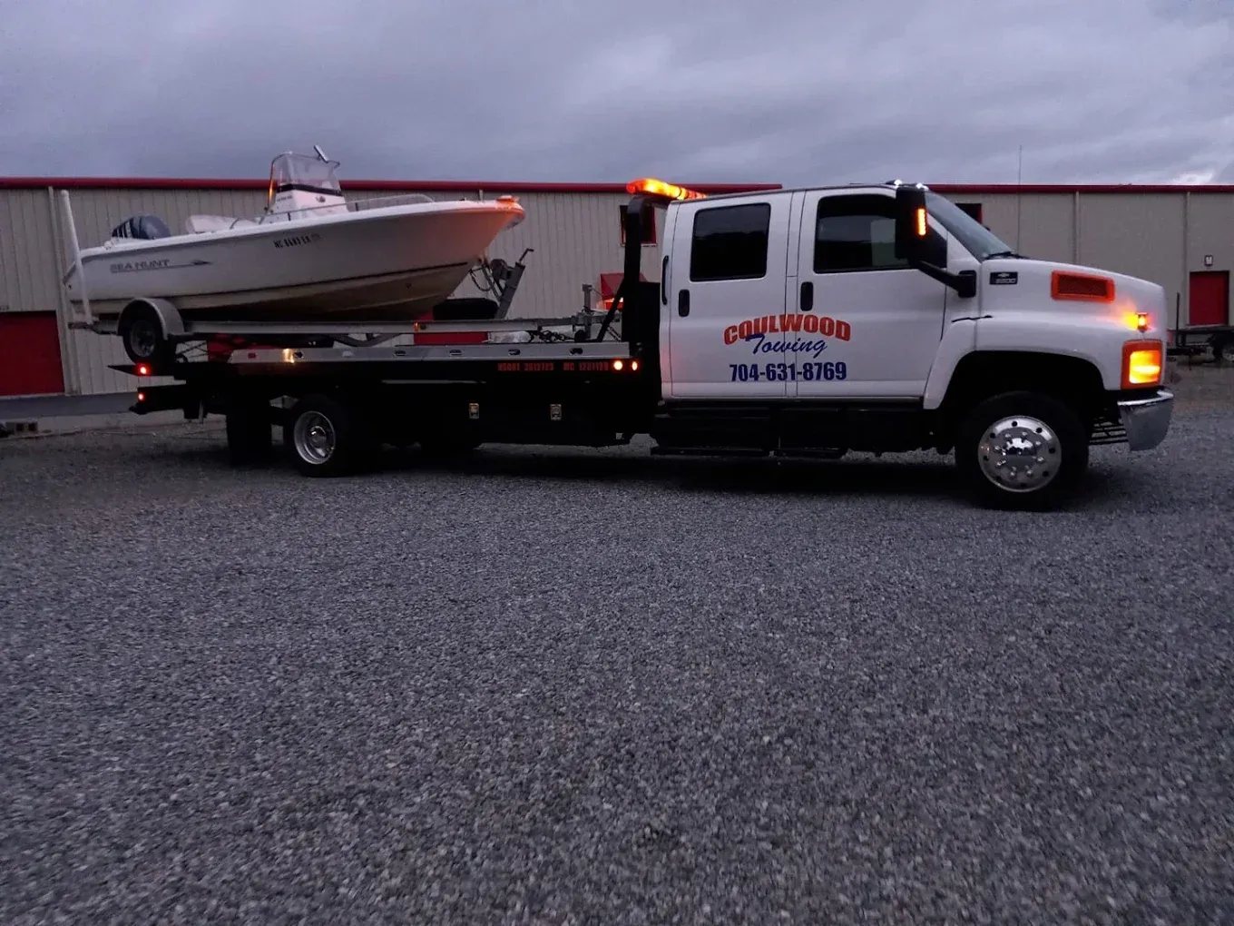 A white flatbed tow truck parked on a gravel lot carrying a white motorboat on a trailer.