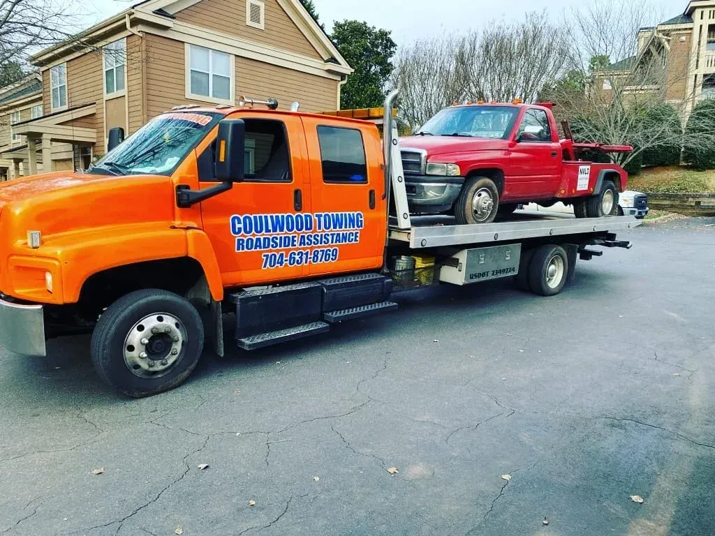 An orange flatbed tow truck carrying a red pickup truck parked on an asphalt lot in front of a tan residential building.