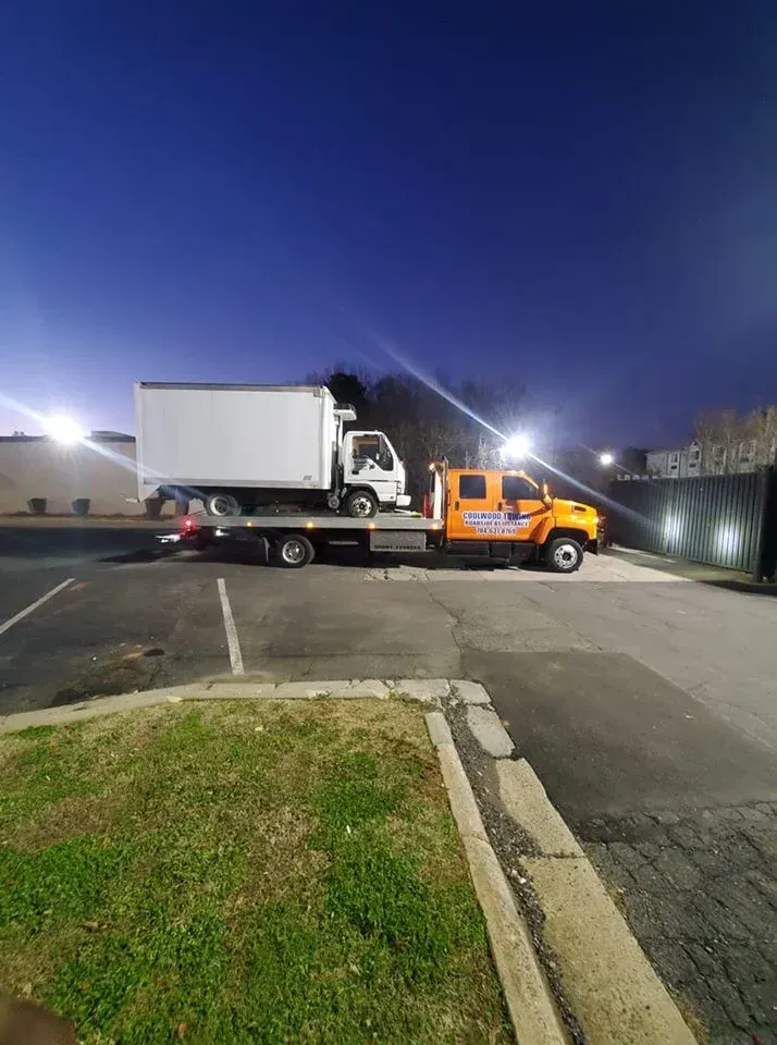 An orange flatbed tow truck carries a white box truck in a parking lot at twilight.
