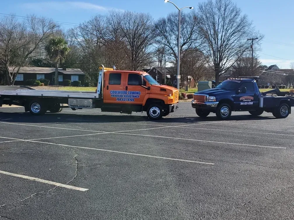 An orange flatbed tow truck and a dark blue truck parked side-by-side in an outdoor asphalt parking lot.
