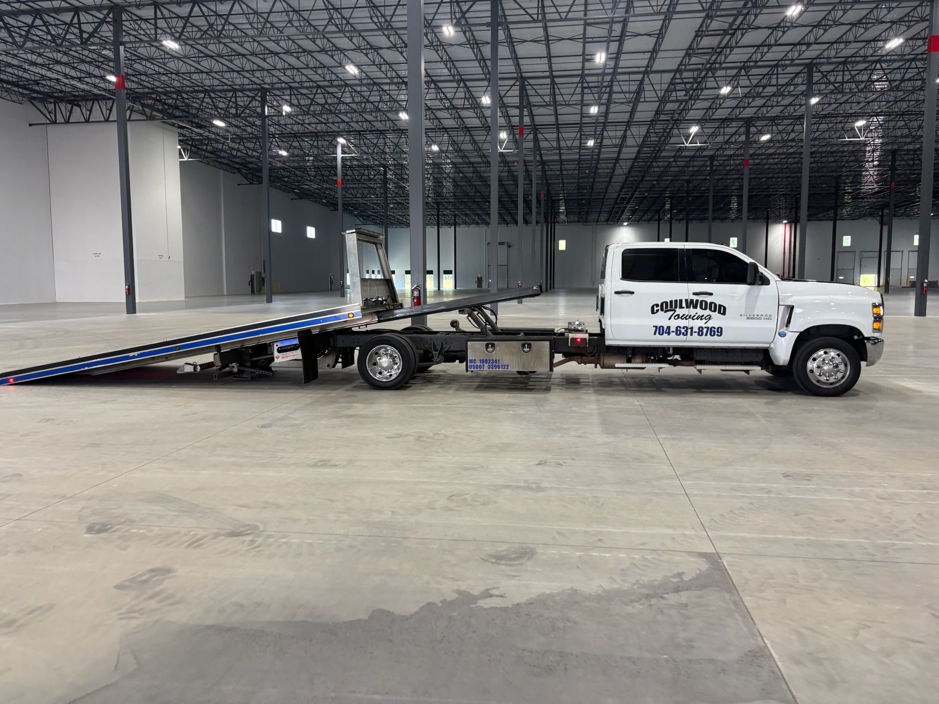 A white flatbed tow truck parked on the concrete floor inside a large, empty industrial warehouse.