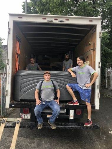 People posing in the back of an open moving truck with furniture inside, parked on a wet street.