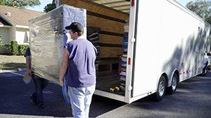 Two people unloading a large wrapped item from a white moving trailer in a driveway