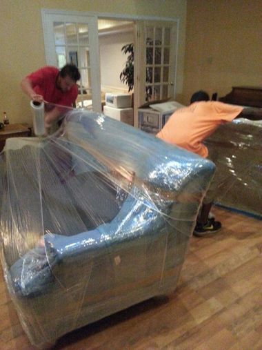 Two people wrapping a couch in clear plastic for moving in a living room