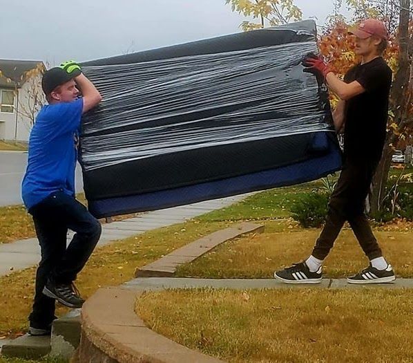 Two people carrying a large black wrapped object outdoors on a sidewalk.