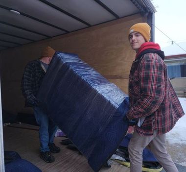Two people carrying a large blue-wrapped mattress into a truck