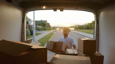 Person unloading cardboard boxes from a van at sunset in a suburban driveway