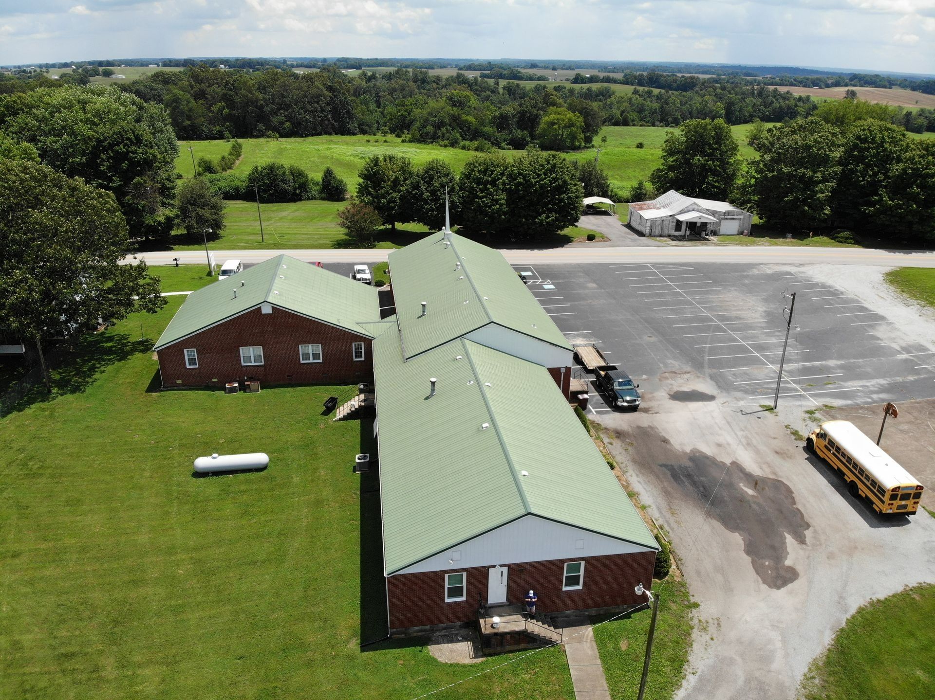 Aerial view of a red brick building with a green roof beside a parking lot and school bus, surrounded by fields.