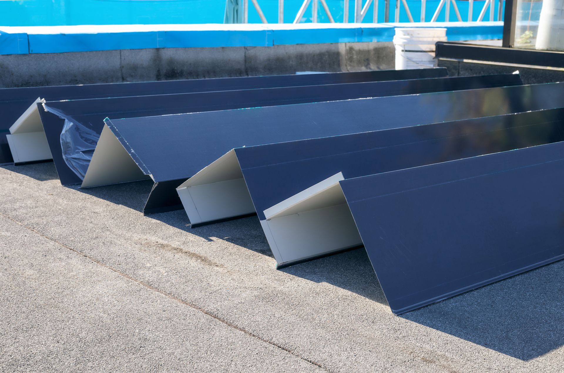 Blue metal roof panels stacked outdoors on a rooftop with a gravel surface and blue tarp in the background