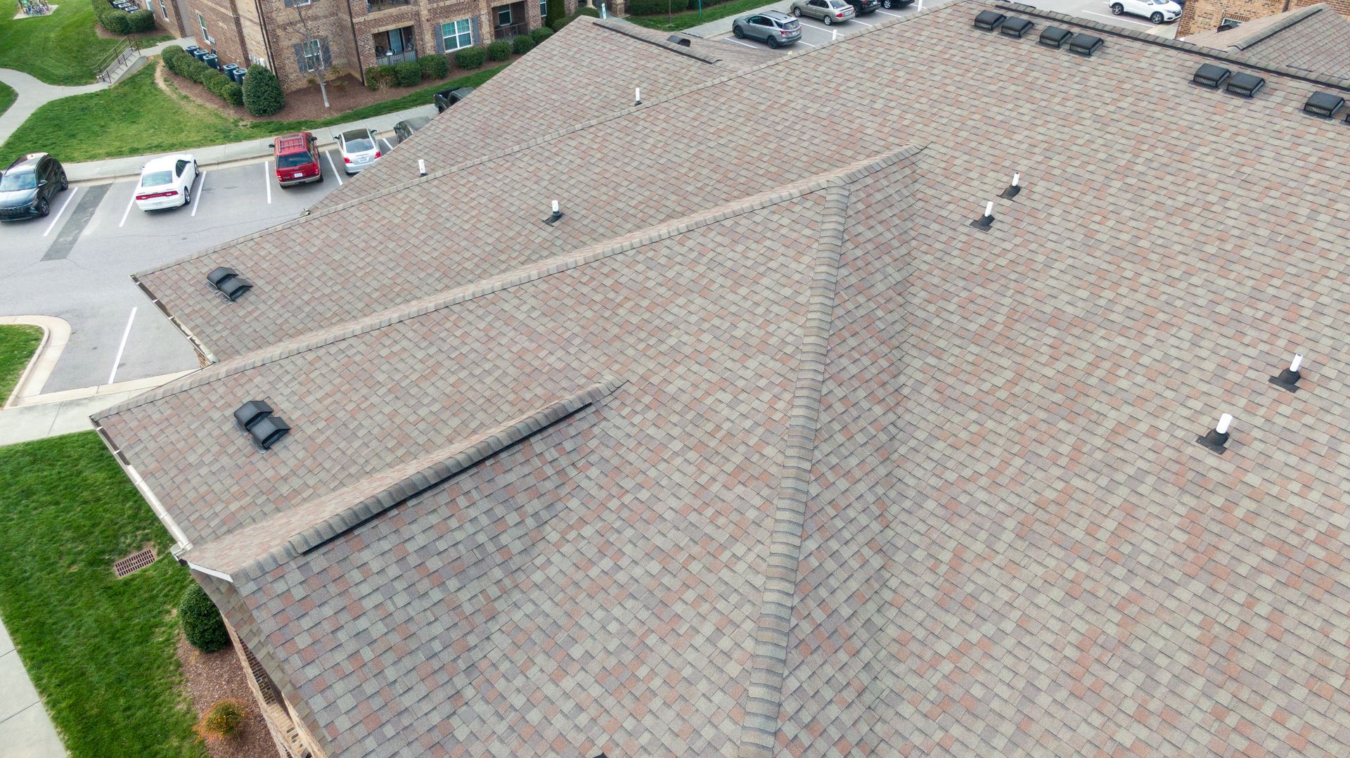 Aerial view of a gray shingle roof with vents beside a grassy parking lot and buildings