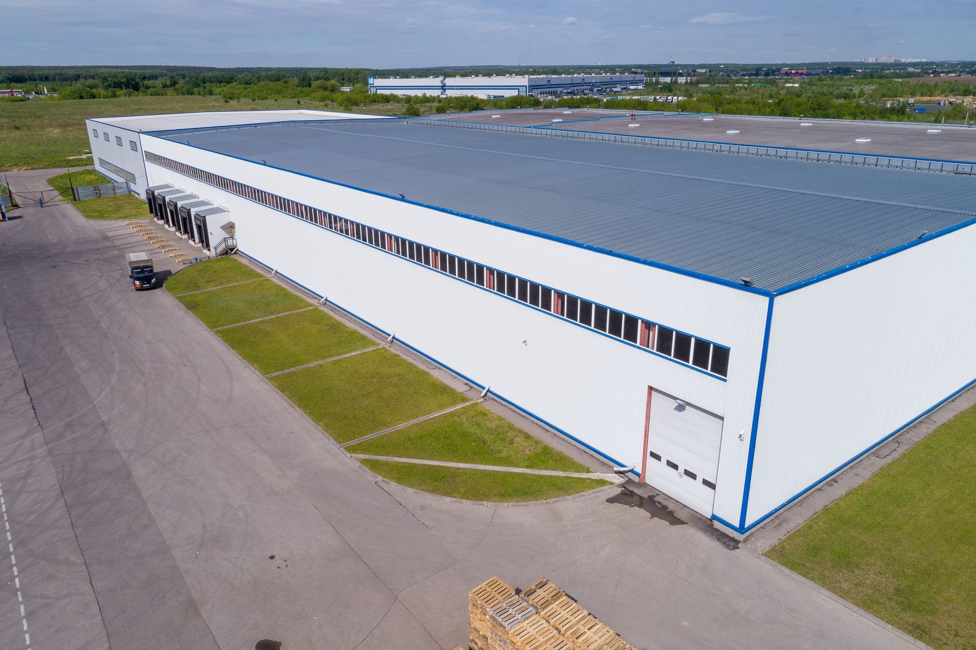 Aerial view of a large white warehouse with blue roof trim beside paved lots and green grass.