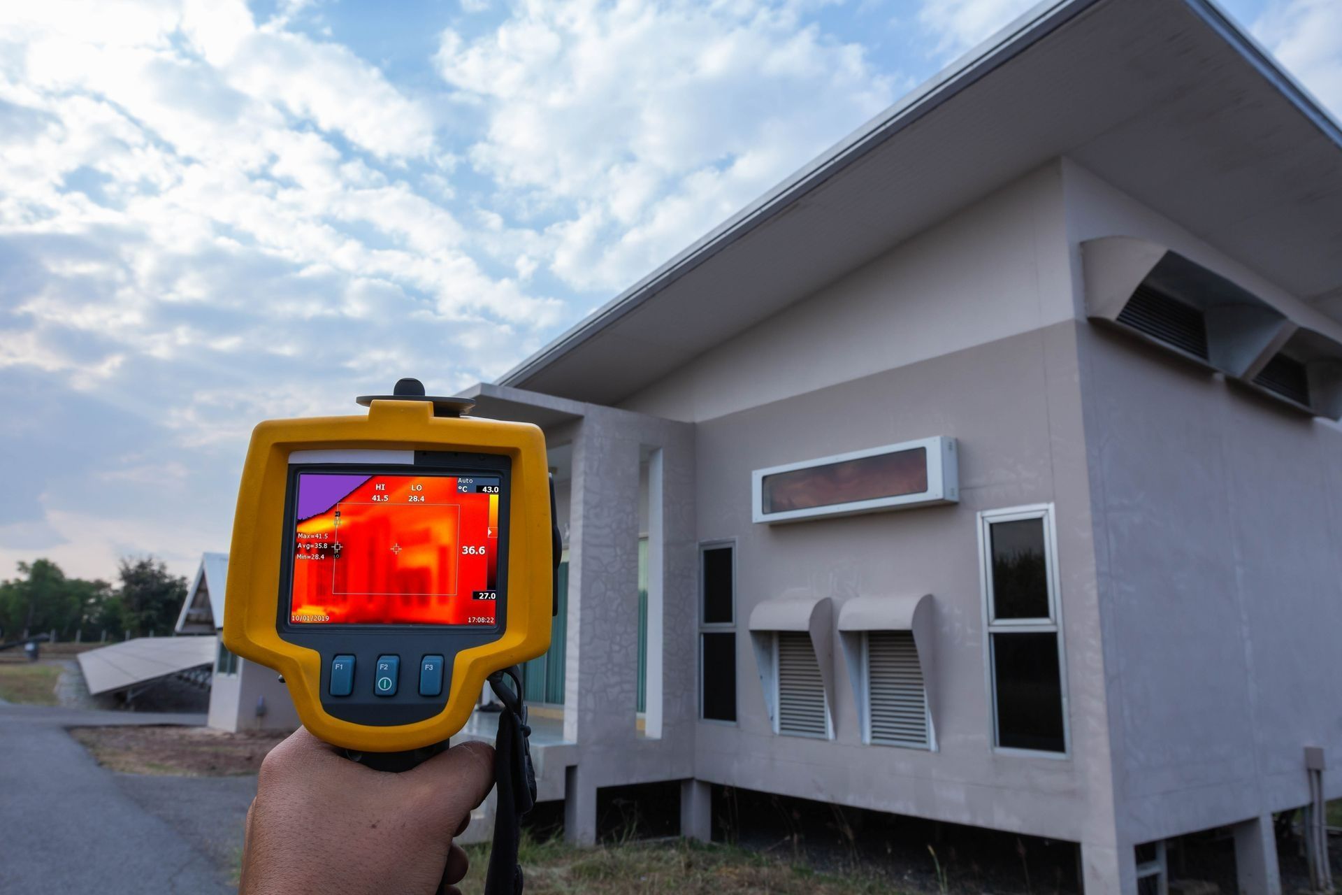 Handheld thermal camera aimed at a house exterior, showing heat signatures in the window and walls.