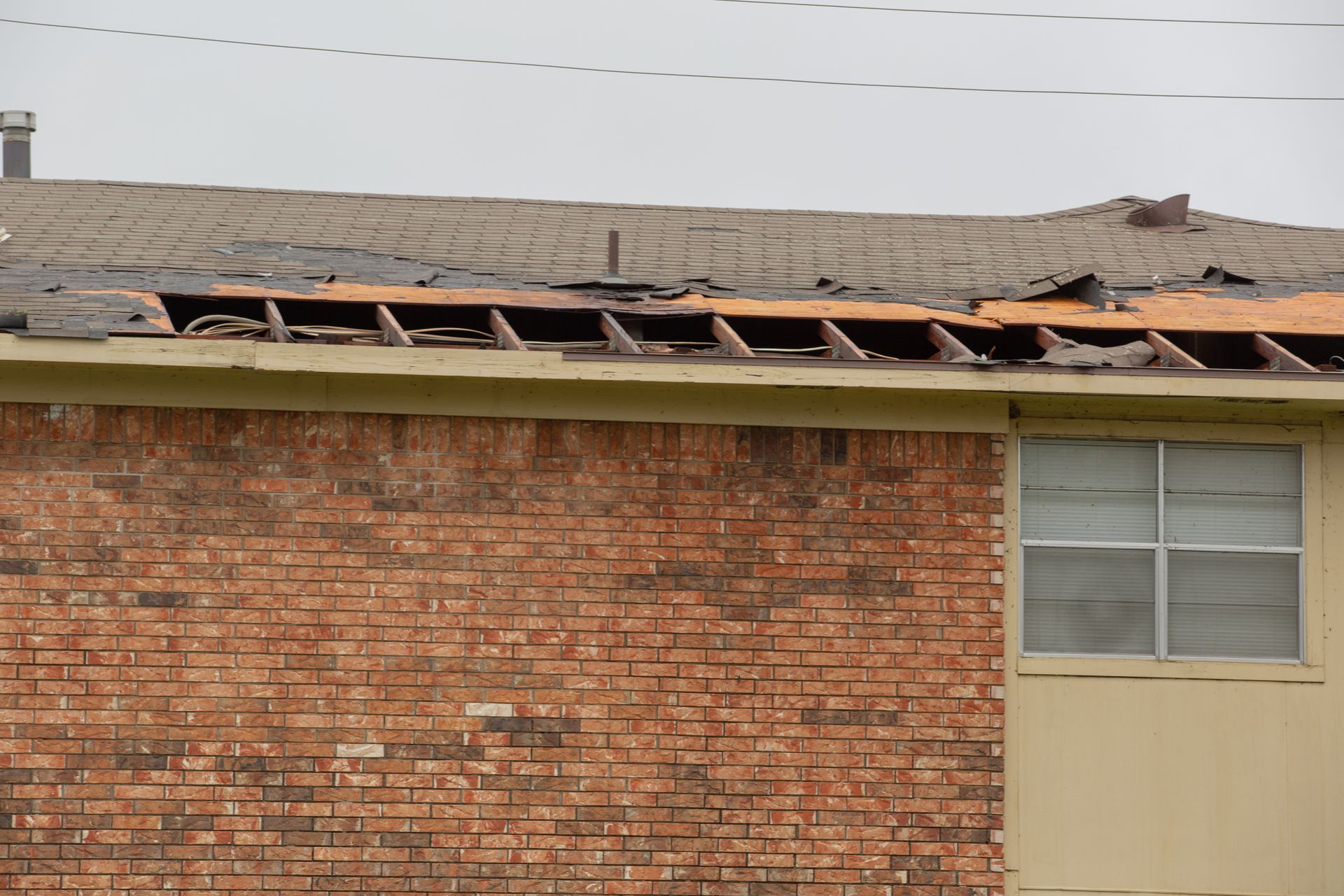 Damaged brick building roof with missing tiles and exposed rafters above a window