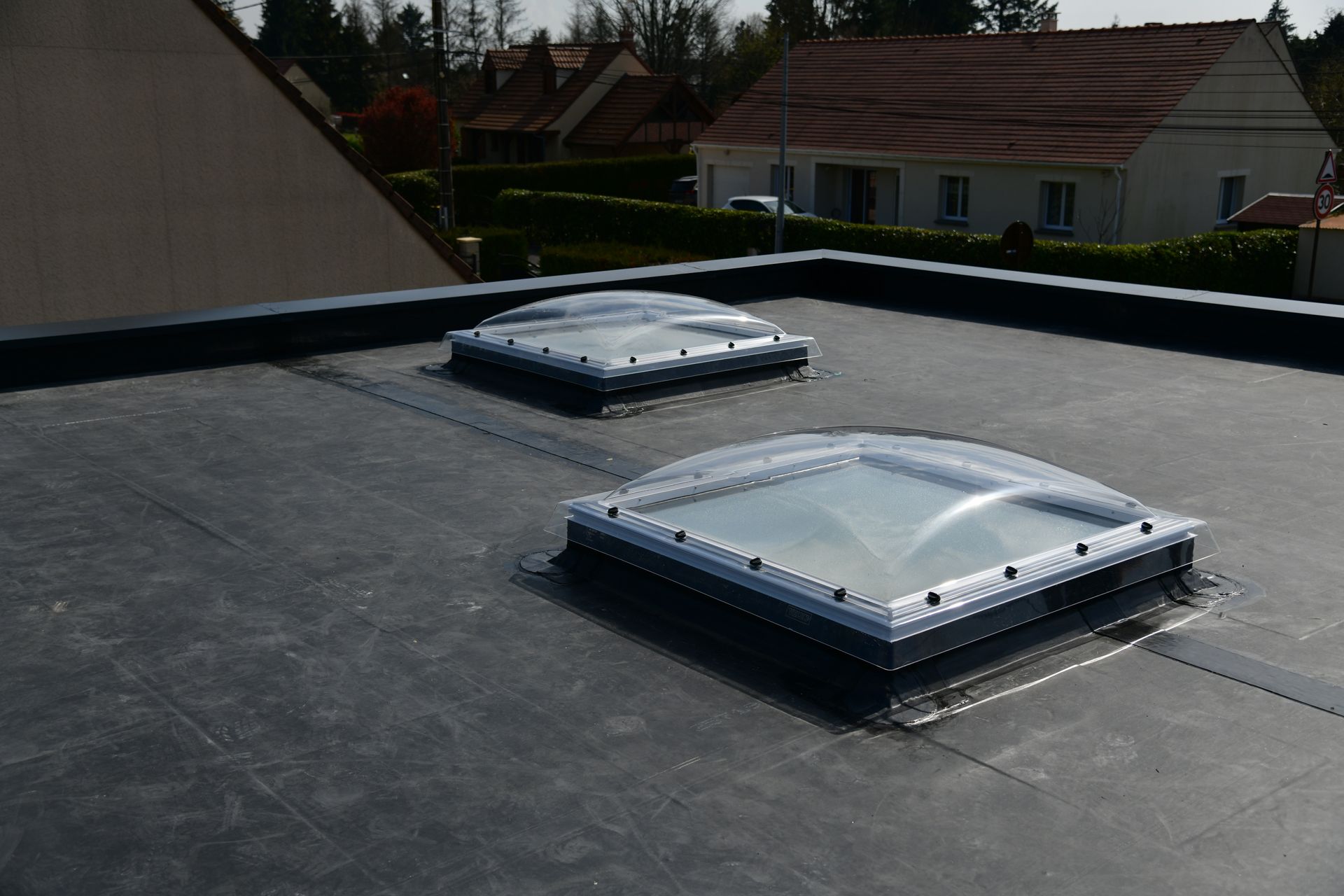 Flat rooftop with two rectangular skylights and a low parapet wall, with houses in the background