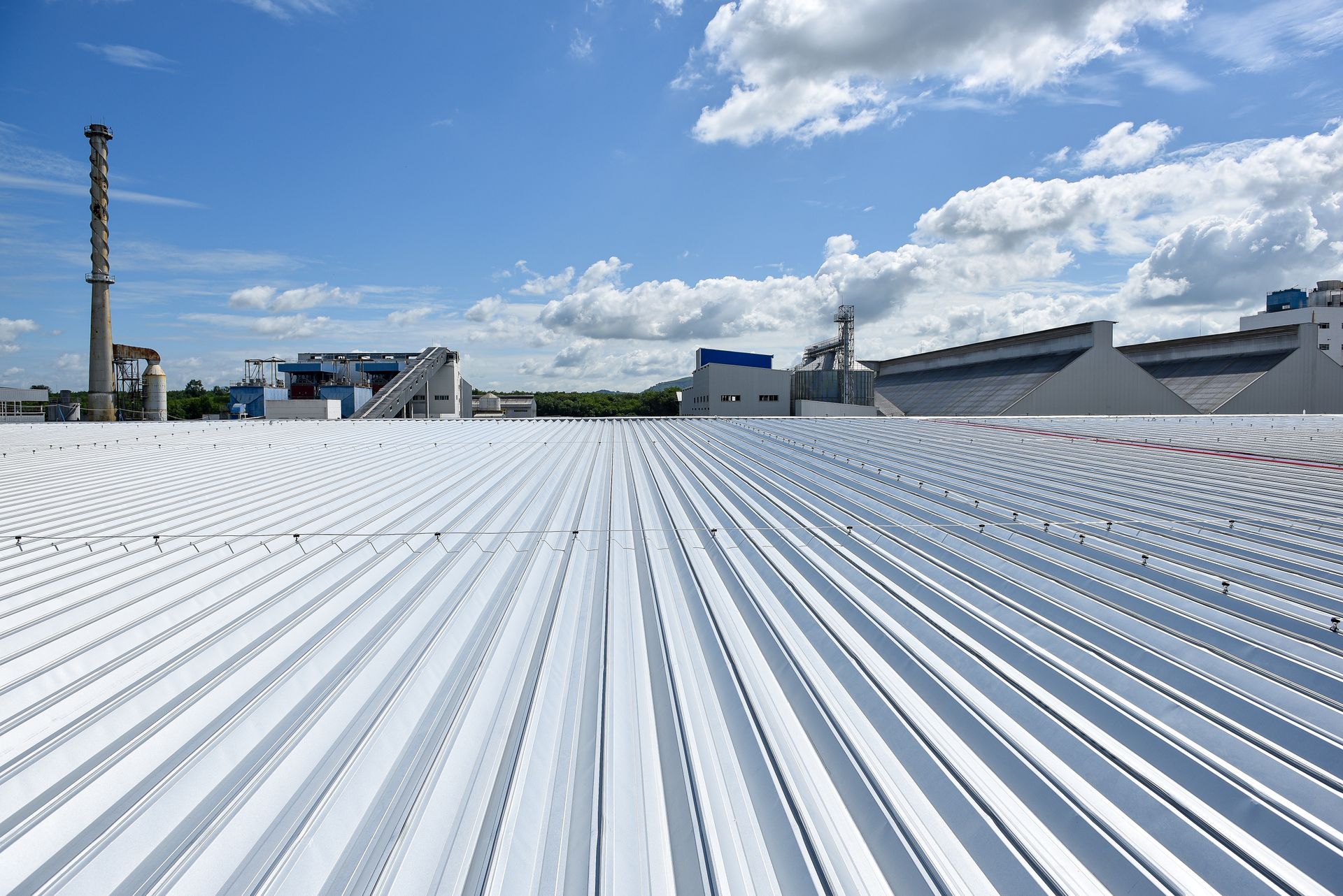 White corrugated metal rooftop under a blue sky with scattered clouds, industrial buildings in the distance