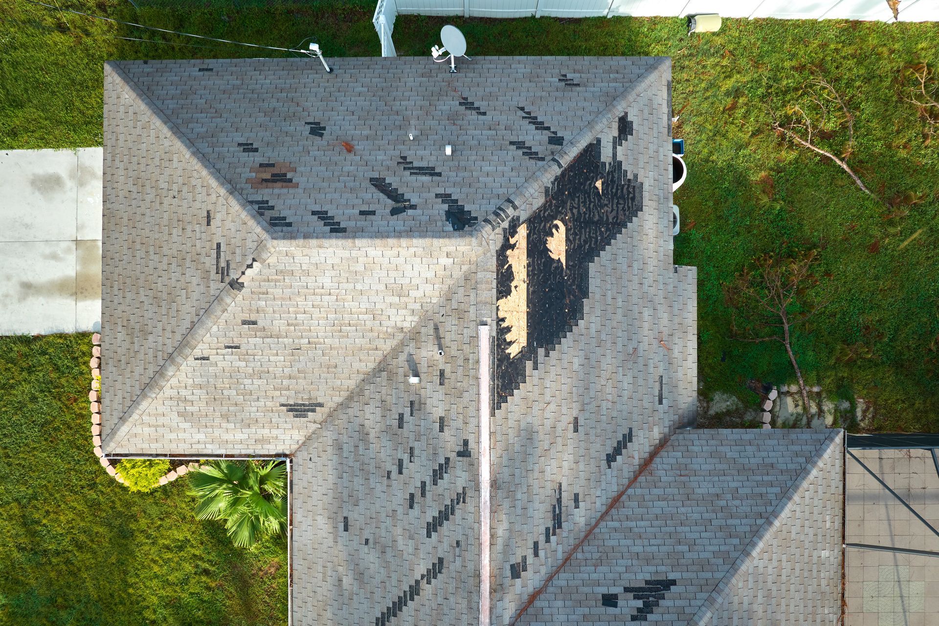 Aerial view of a gray house roof with weathered shingles beside green grass.