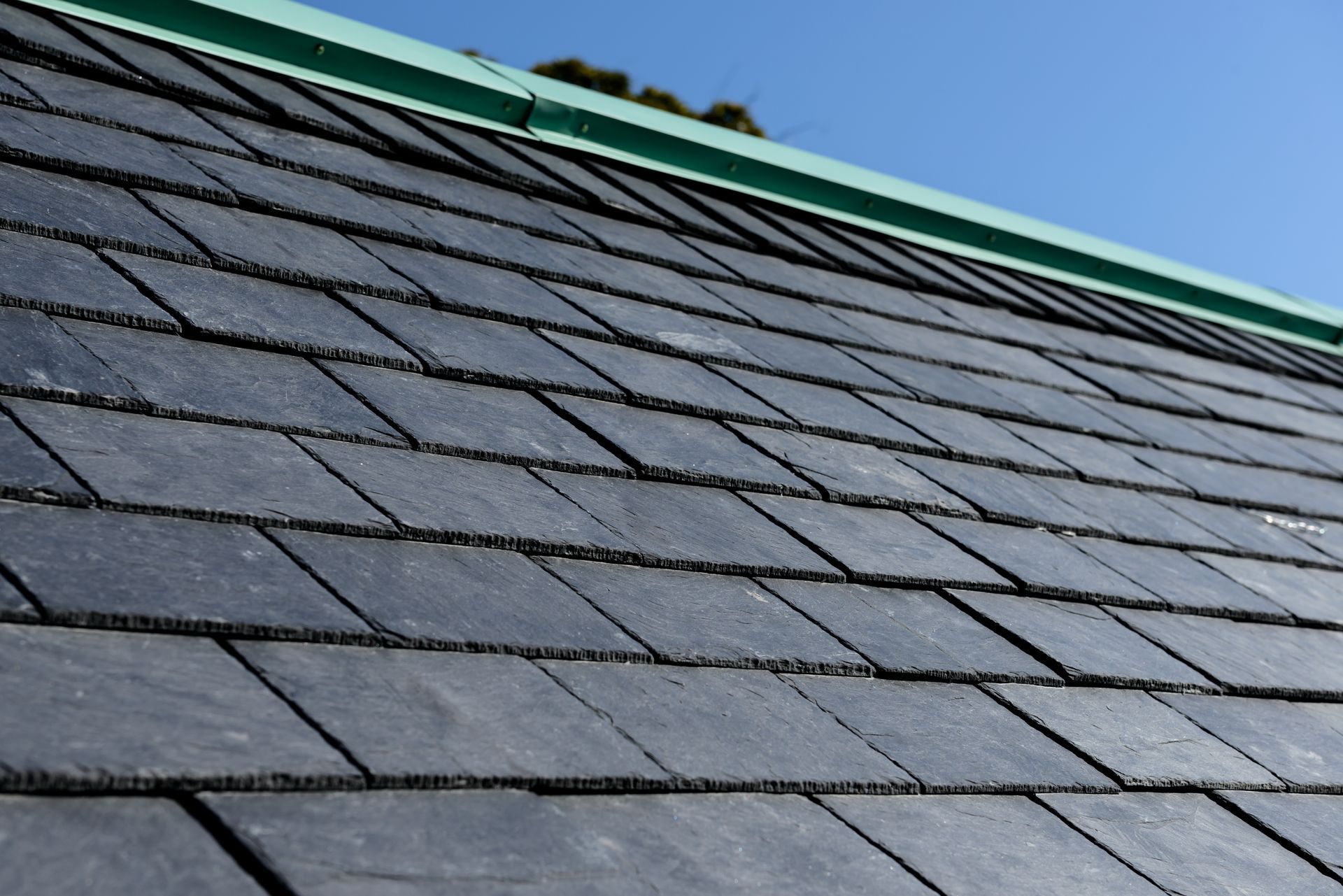 Close-up of a dark gray shingled roof with a green trim against a blue sky