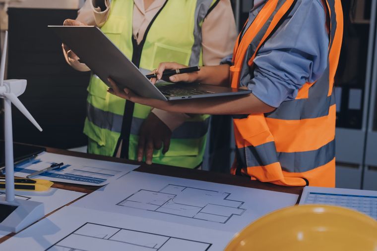 Two workers in orange safety vests review blueprints on a laptop at a construction site