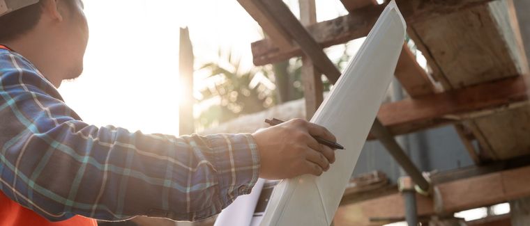 Person in a plaid shirt holding rolled-up plans at a construction site in warm sunlight