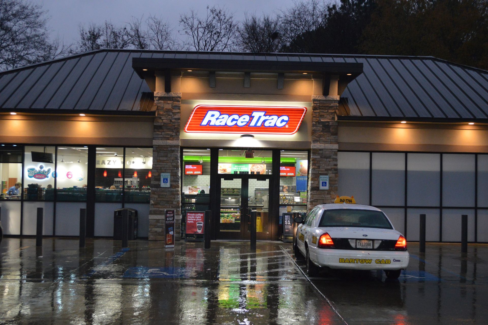 RaceTrac gas station storefront at dusk, wet pavement reflecting lights, silver car parked outside.