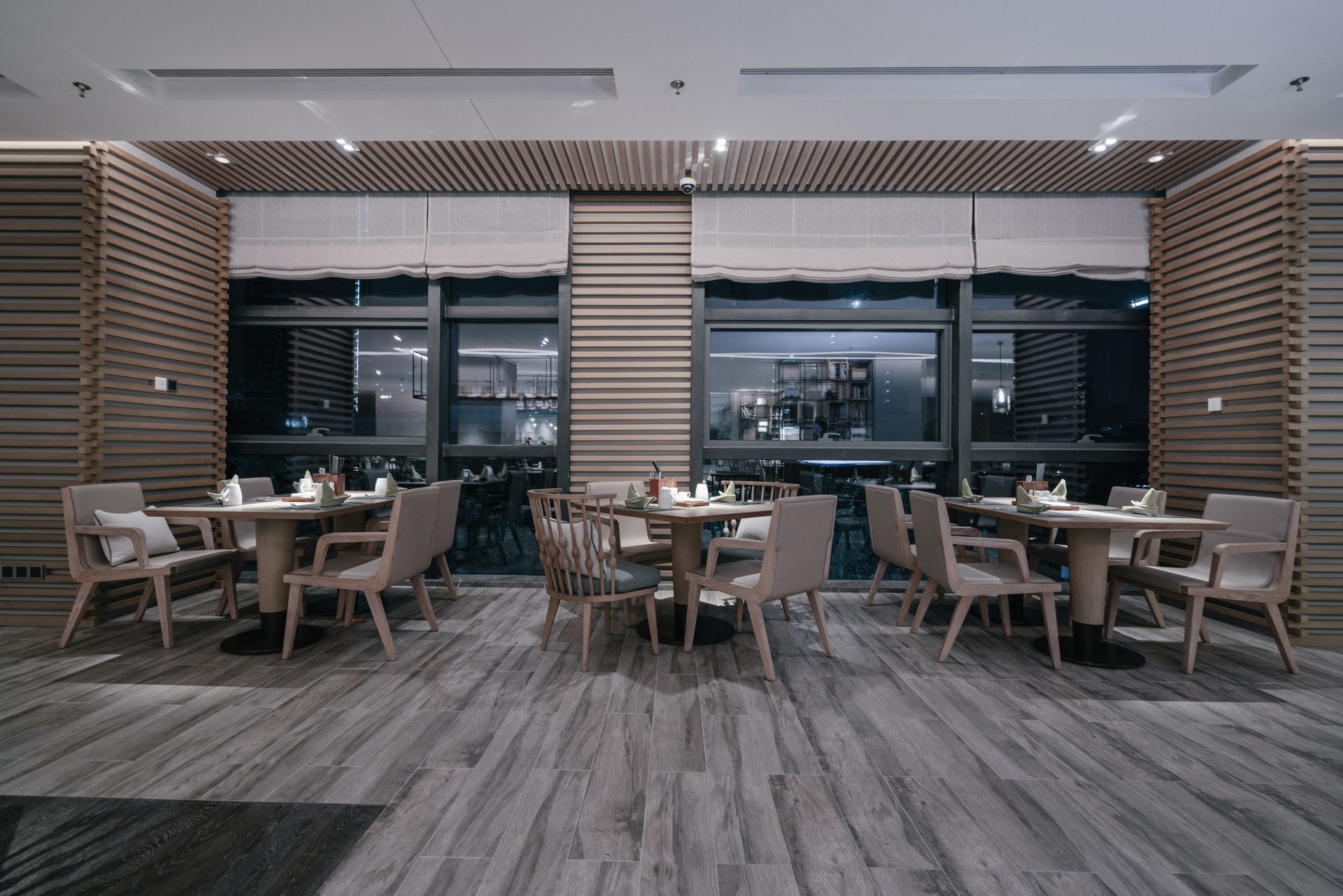 Modern restaurant dining area with empty tables, chairs, wood-paneled walls, and large windows at night