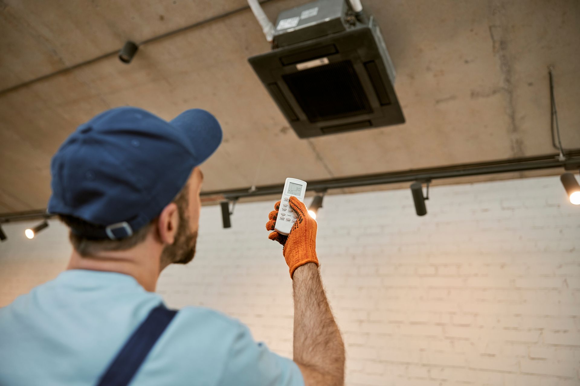 Person in a blue cap points a remote at a ceiling-mounted unit in a room with a brick wall.