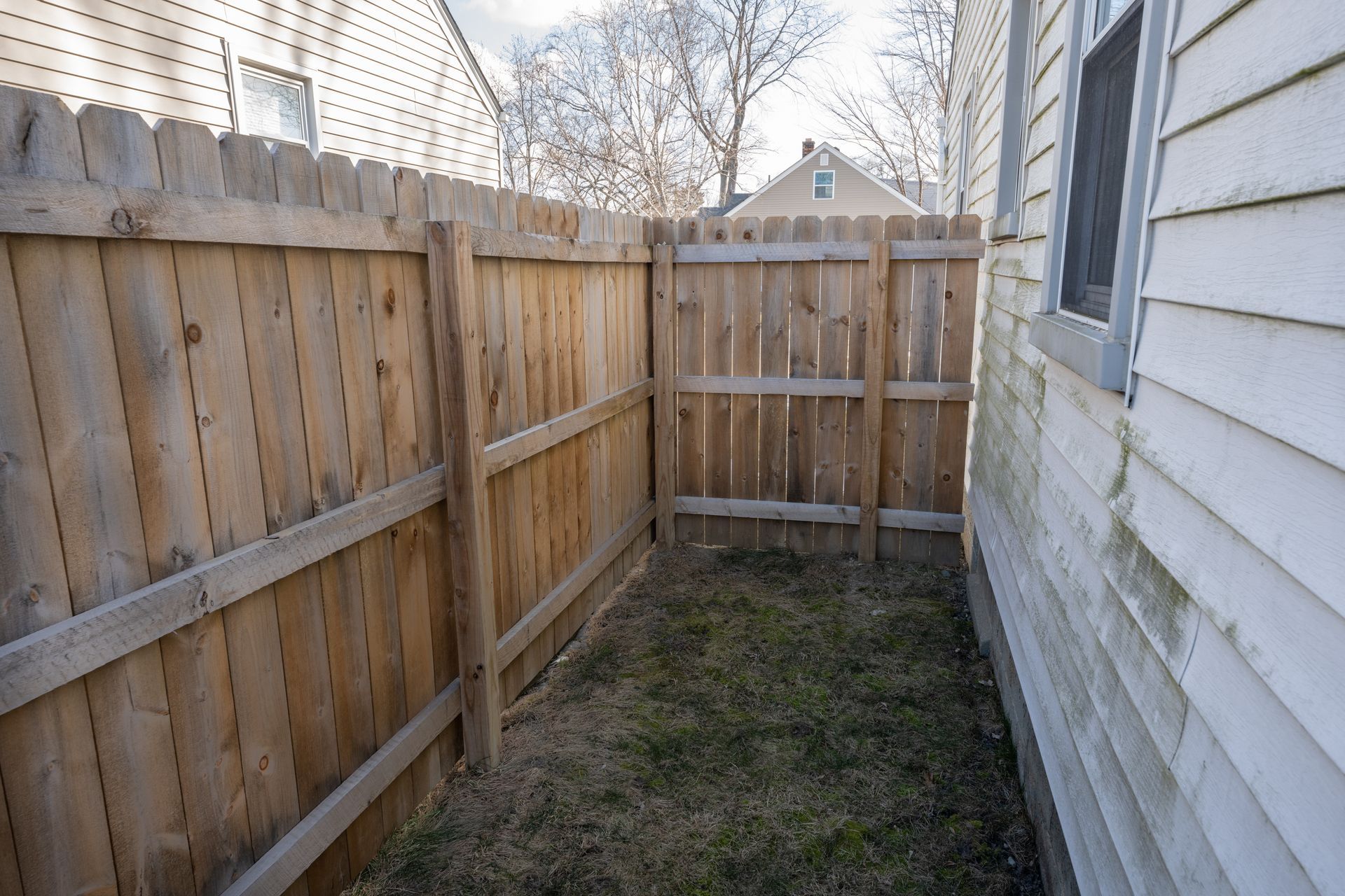 Narrow backyard space with wooden privacy fences and the side of a white house, covered in patchy grass.