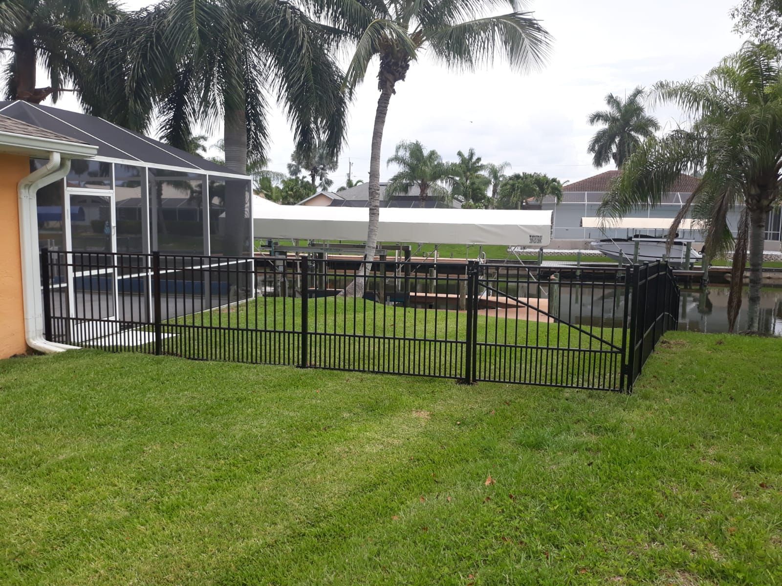 Black fence enclosing a backyard with a screened porch and canal view; green grass and palm trees.