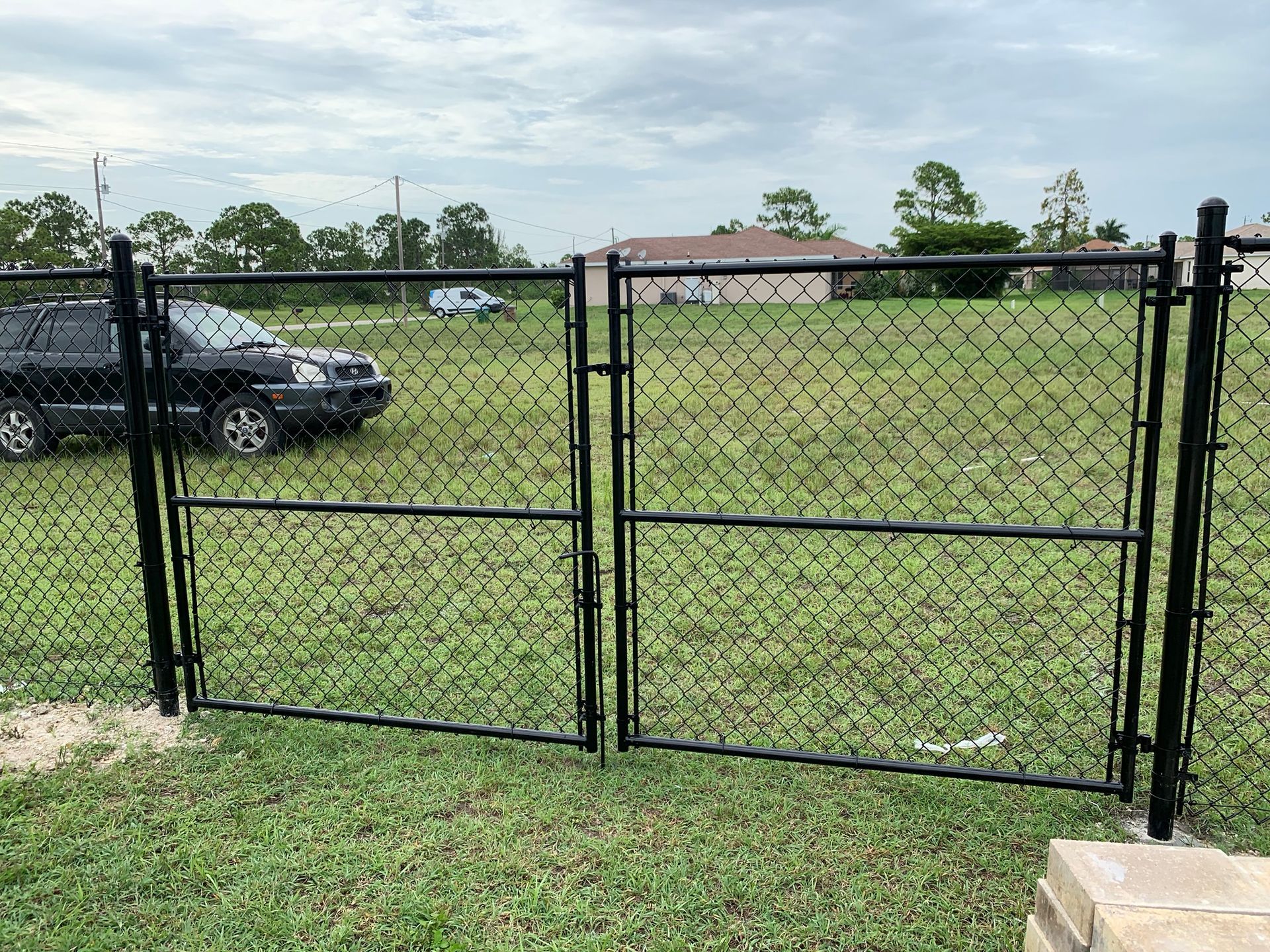 Black chain-link fence with double gates in a grassy area, a dark SUV and houses in the background.