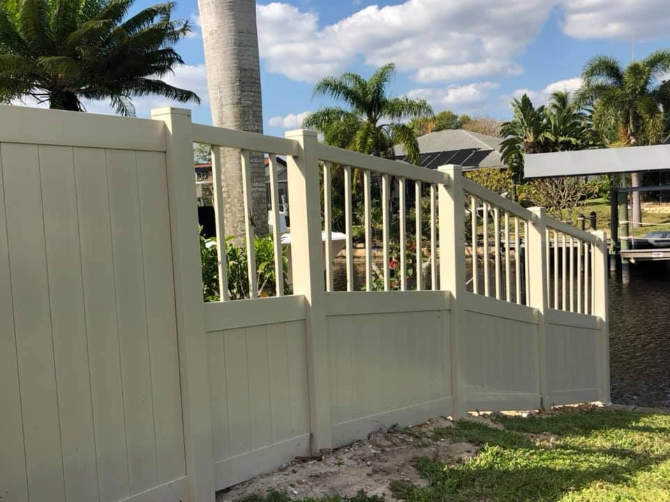 Beige vinyl fence near water with open top section; palm trees and houses in the background.