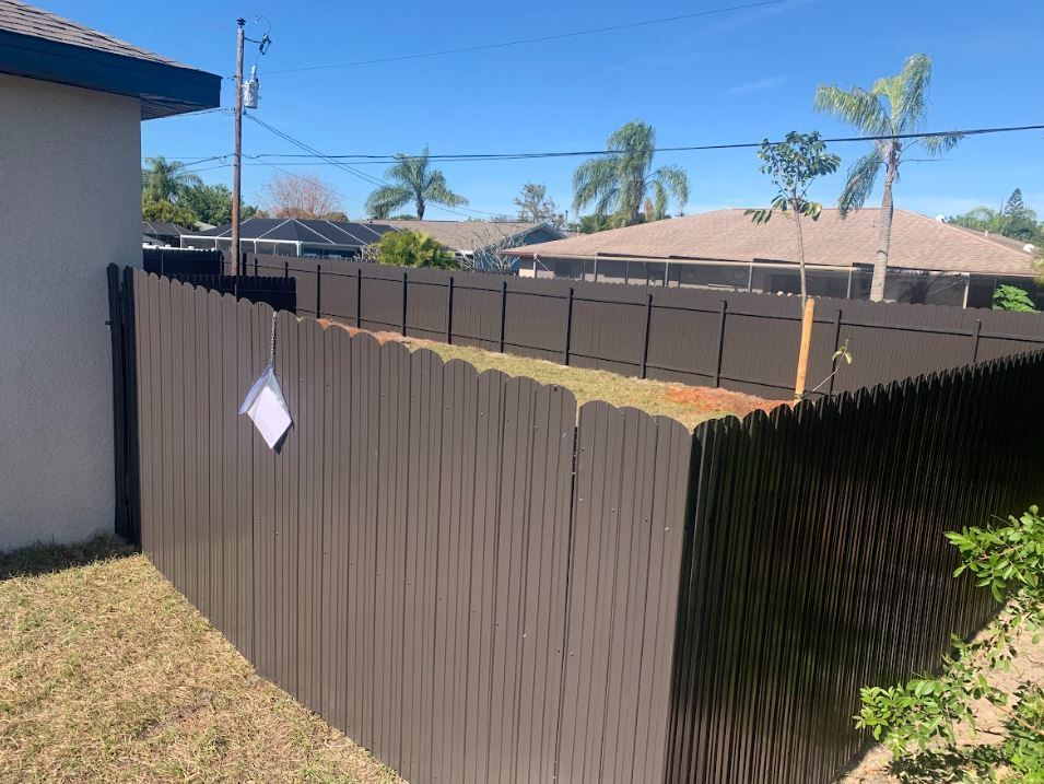 Brown picket fence surrounding a yard, with a light blue sky and some houses visible in the background.
