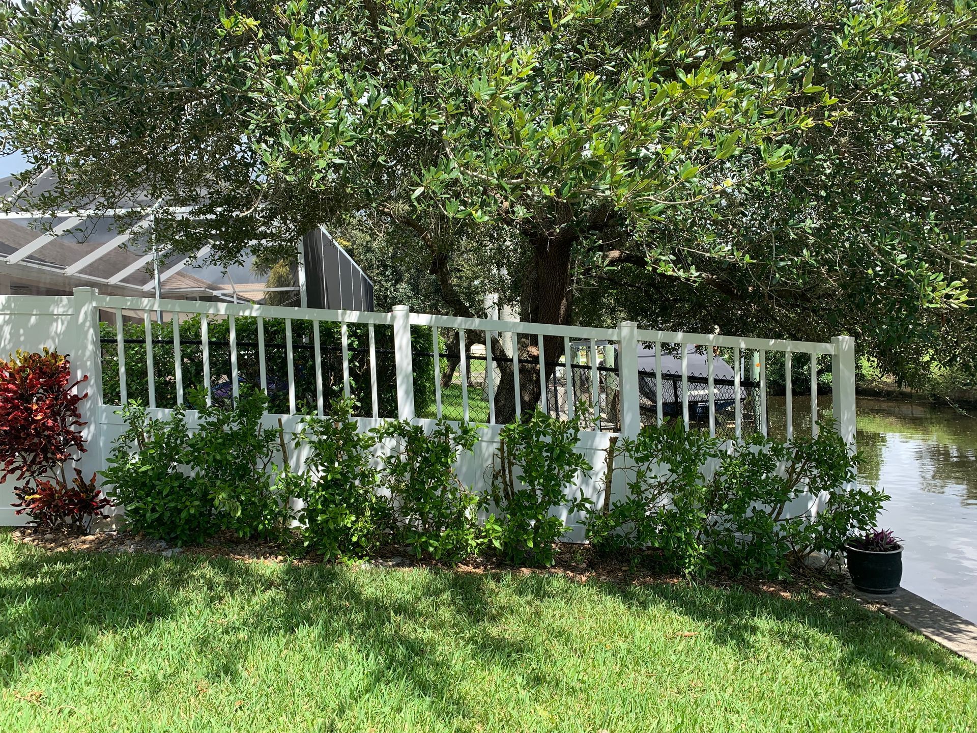 White picket fence with green bushes in front, trees behind, and a canal.