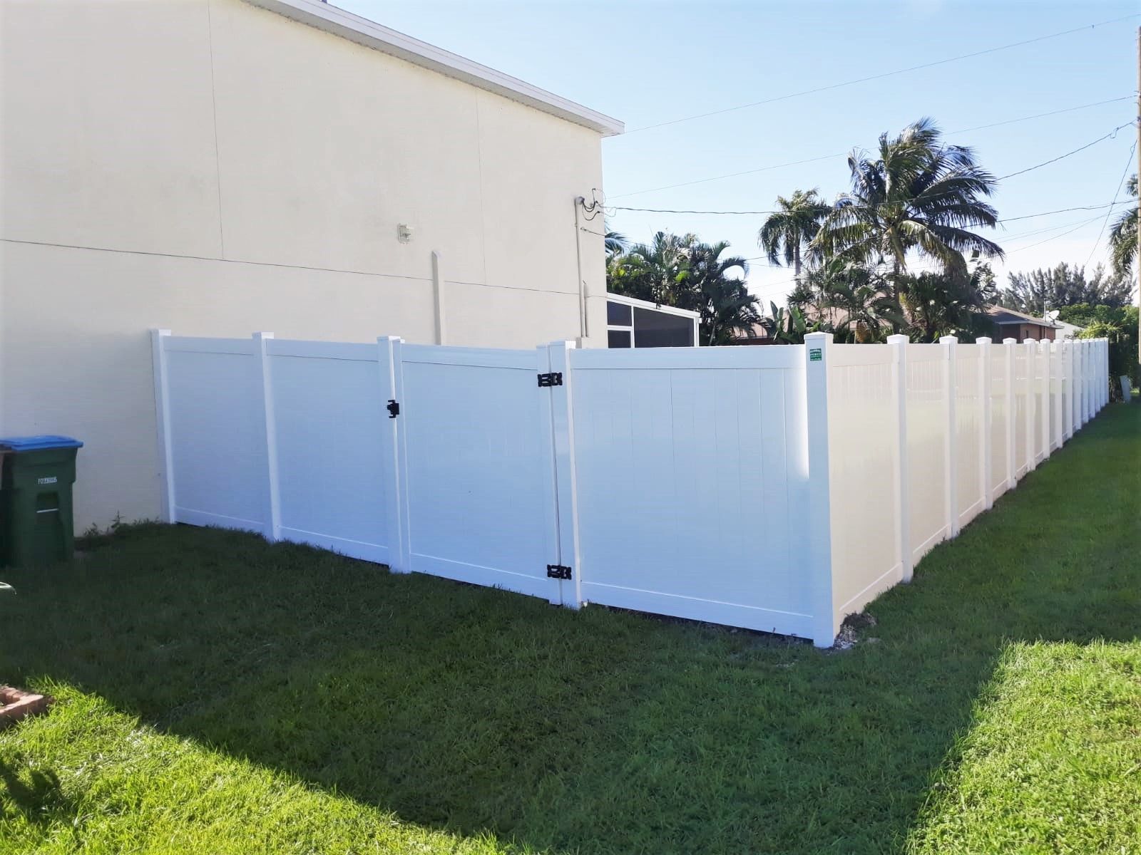 White vinyl fence surrounds a green lawn, adjacent to a beige building, under a sunny sky.