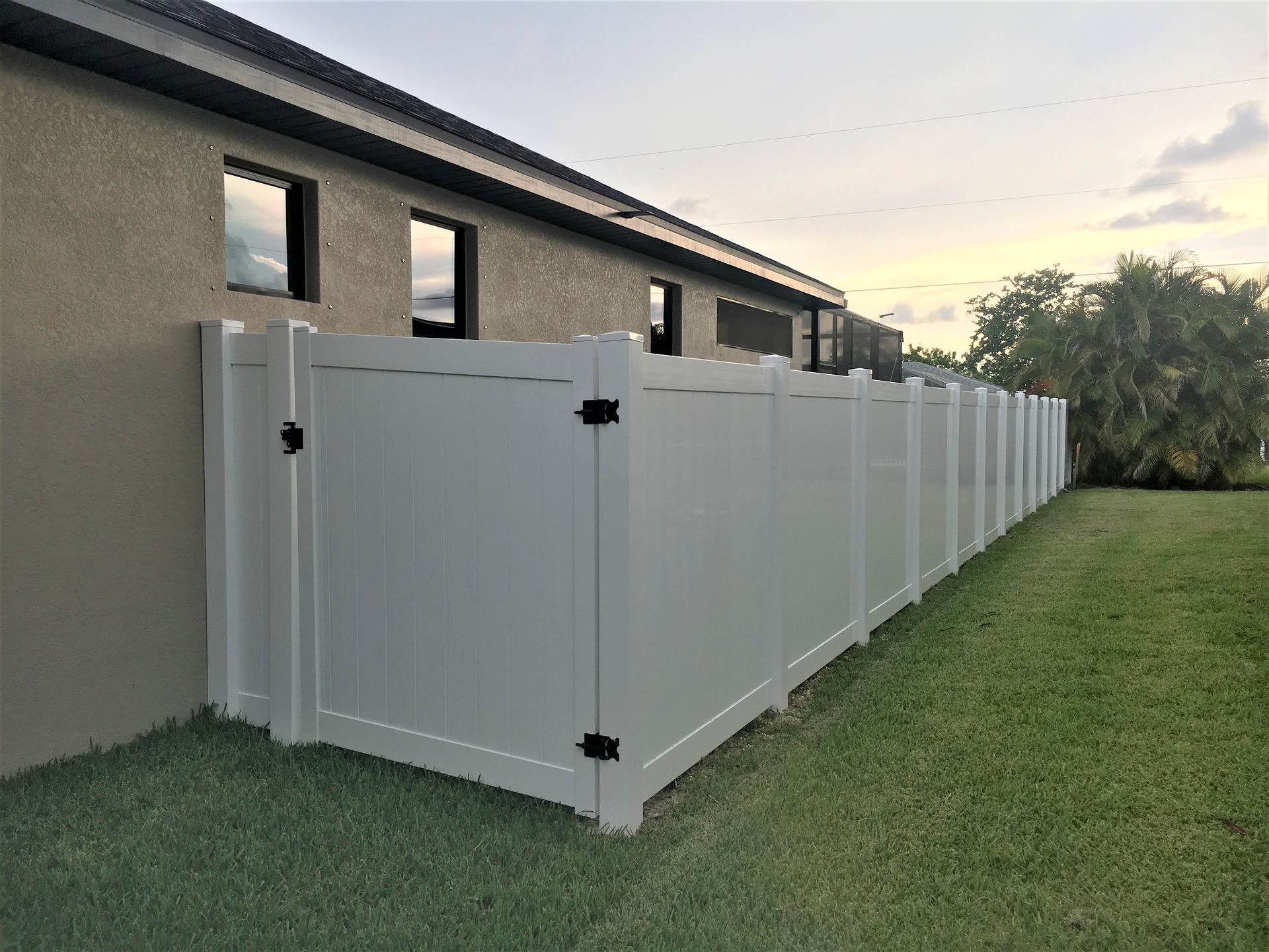 White vinyl fence along a grassy backyard, adjacent to a tan building with dark-framed windows.