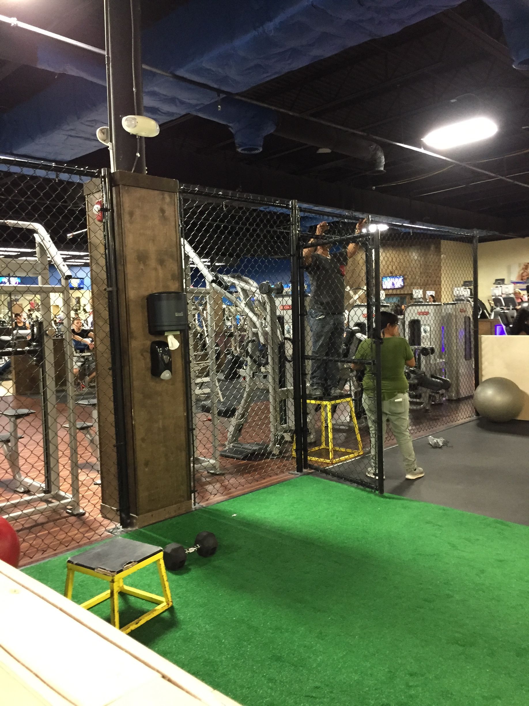 A gym interior featuring a cage-like training area, person near a punching bag, exercise equipment, and green turf.