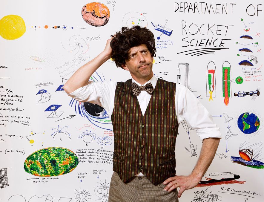 Man in vest posing in front of a rocket science whiteboard with colorful science doodles