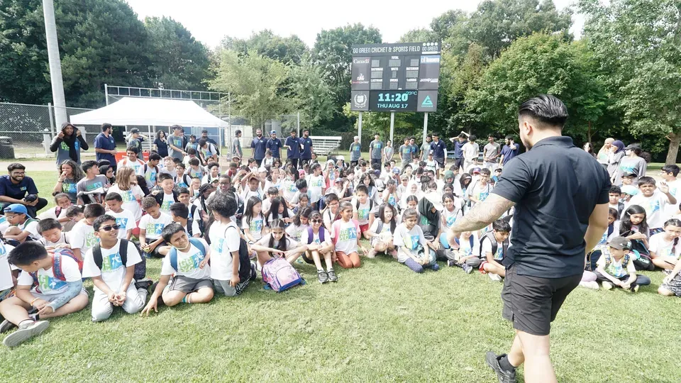 A man is standing in front of a large group of people sitting on the grass.