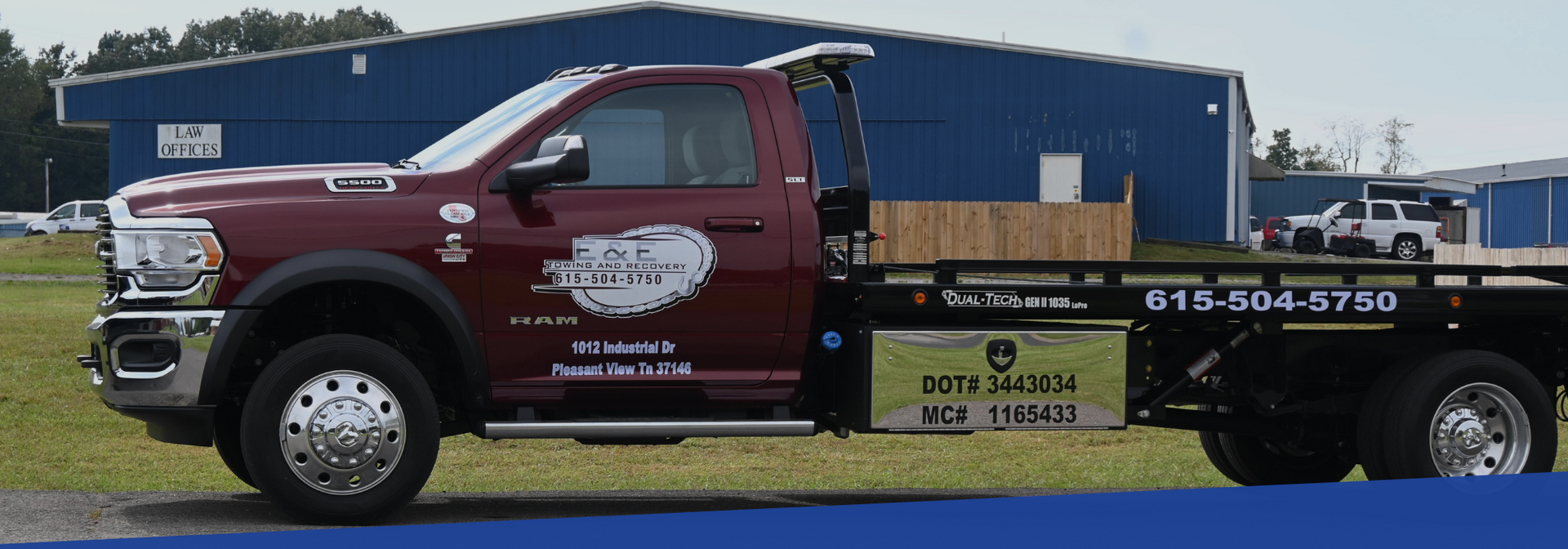 Burgundy tow truck with John Deere logo in front of blue building.
