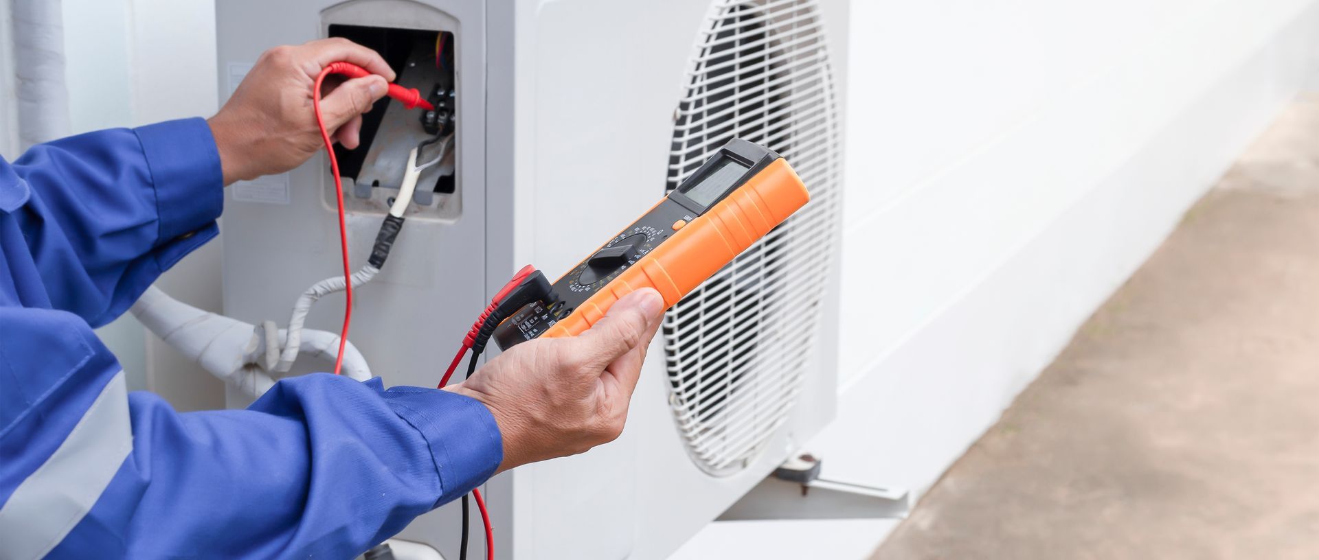 Electrician testing an outlet with a multimeter beside a wall-mounted HVAC unit