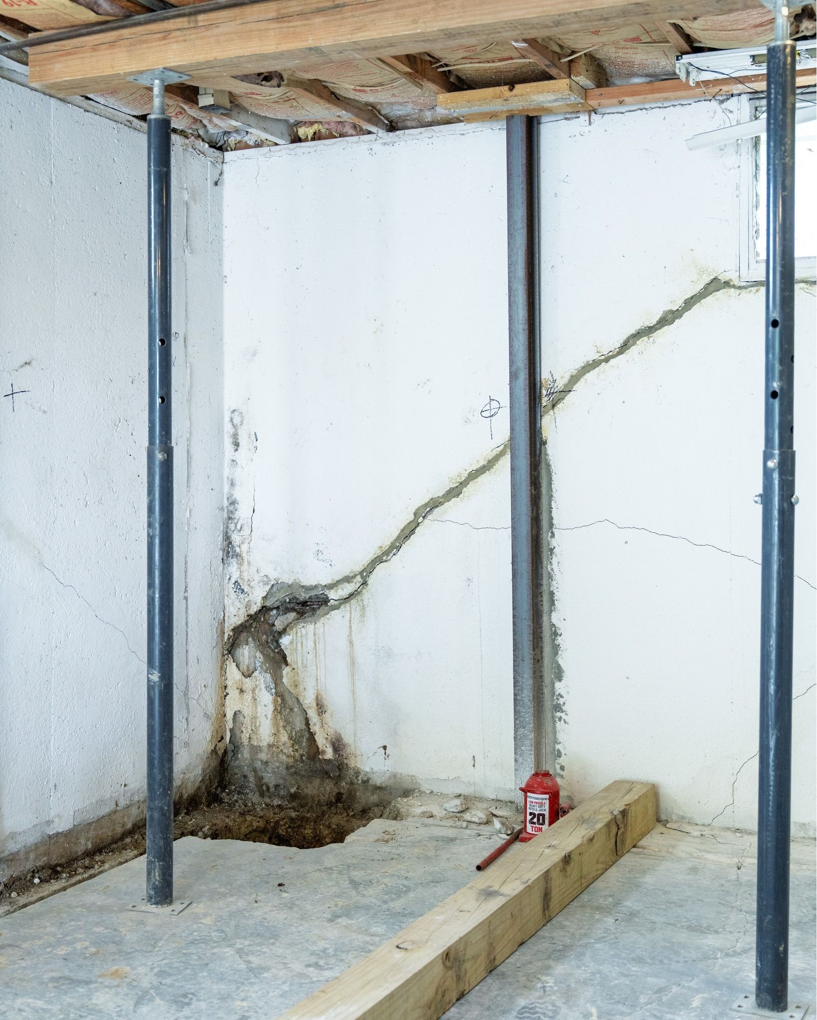 Basement corner with cracked concrete wall, dark water stain, exposed ceiling, and metal support posts