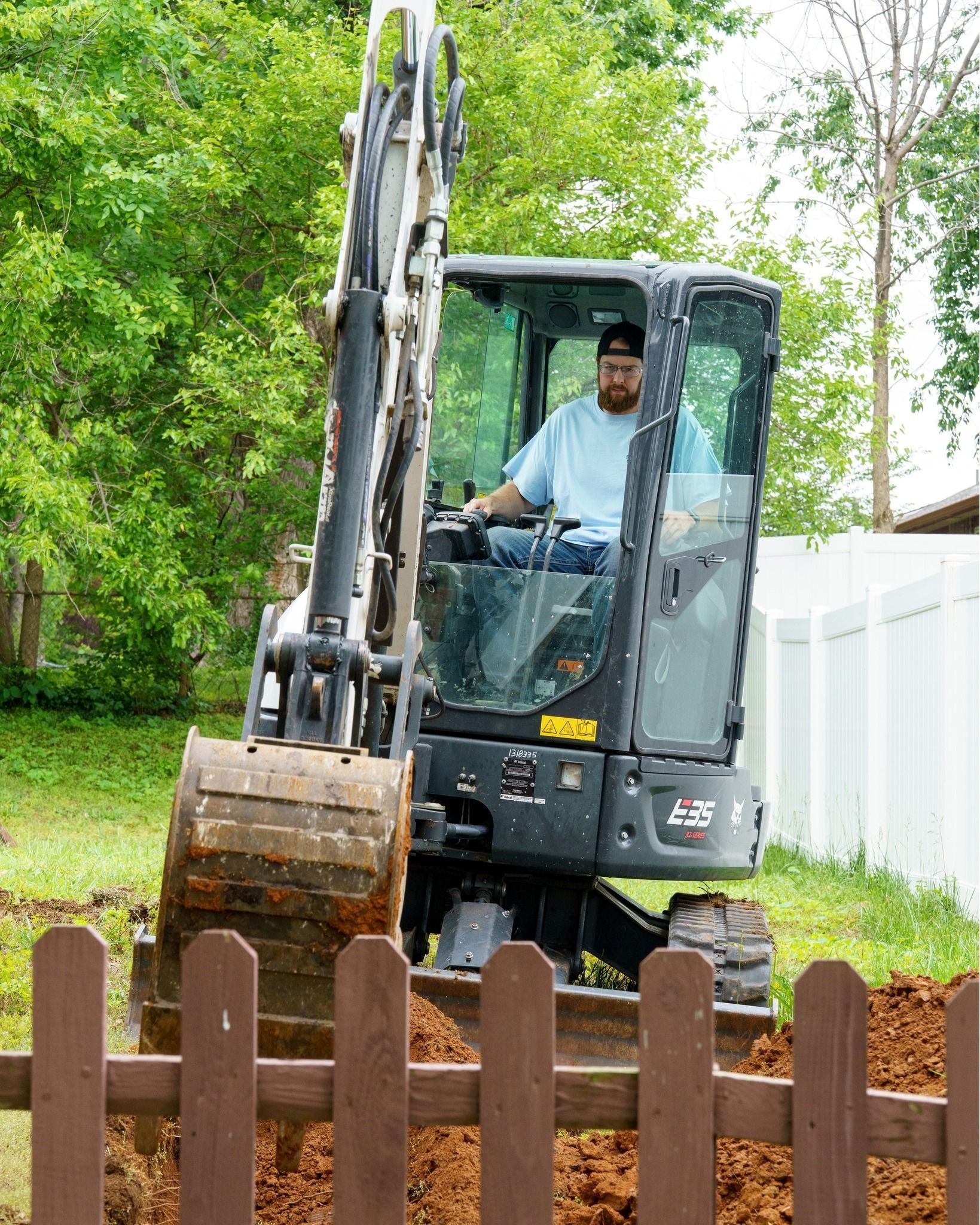 Excavator digging soil in a yard beside a white fence and wooden picket fence, with green trees behind.