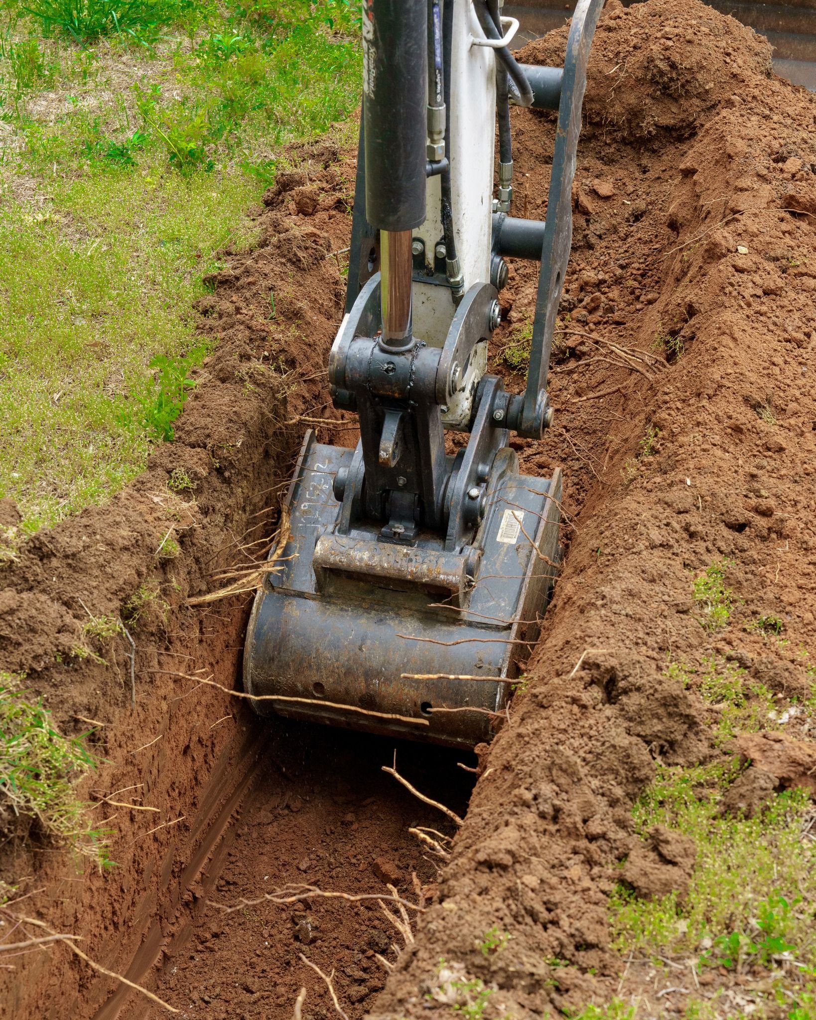 Excavator bucket digging a trench through brown soil beside a grassy patch