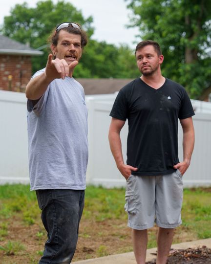 Two men standing outdoors near a house; one points toward the camera while the other looks on.