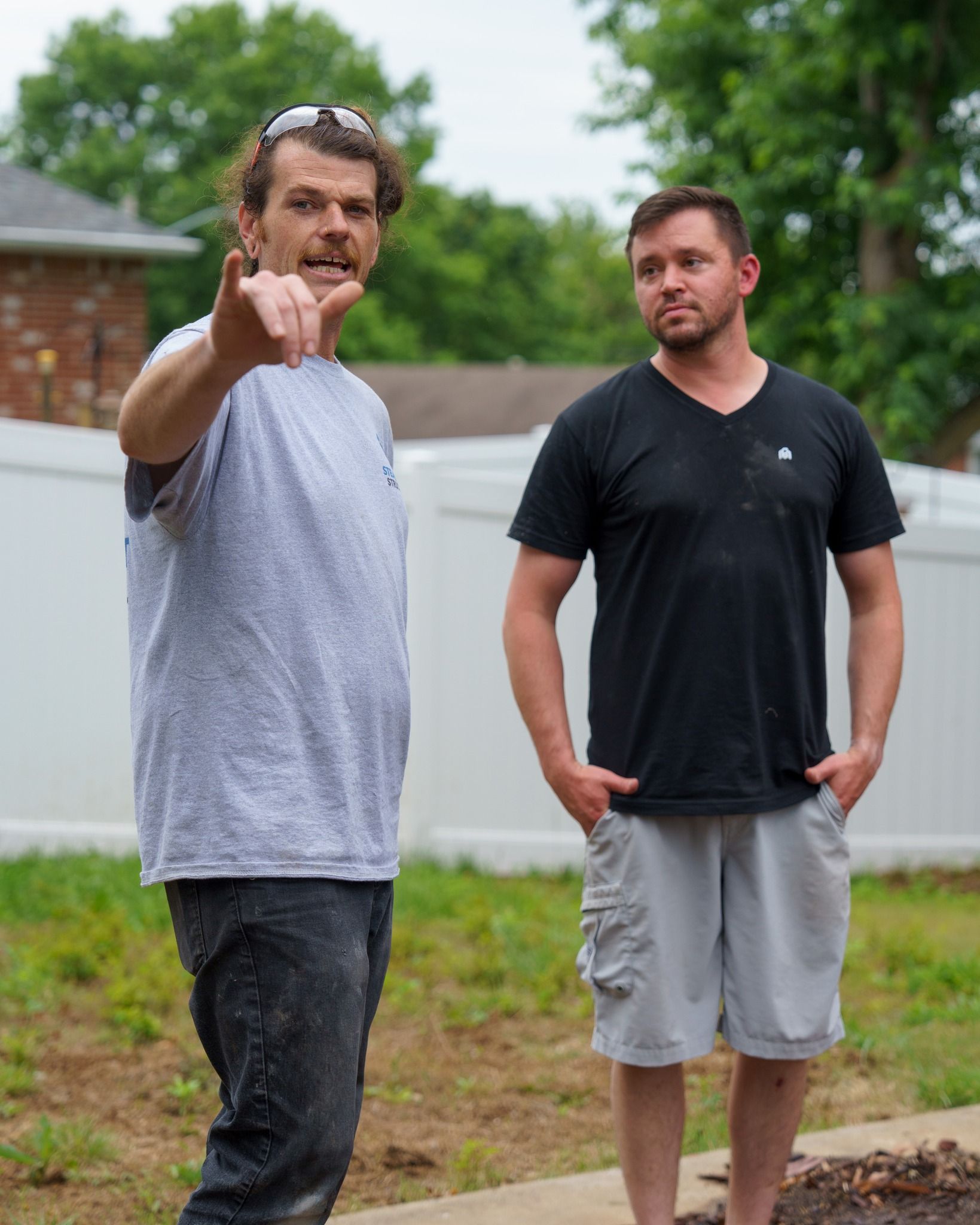 Two men outdoors; one points toward the camera while the other stands nearby in a backyard.