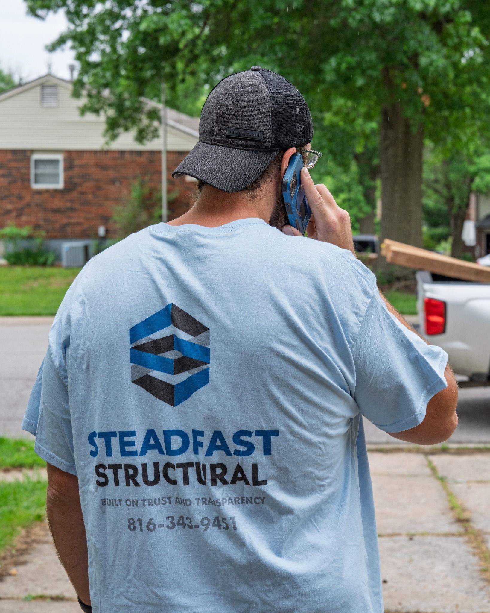 Worker in a light blue Steadfast Structural shirt talking on a phone outdoors near a pickup truck.