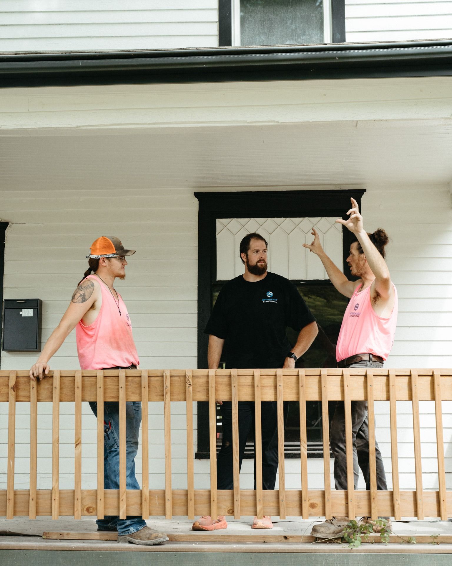 Three people on a porch, two in pink vests gesturing toward a window, one man standing in the doorway